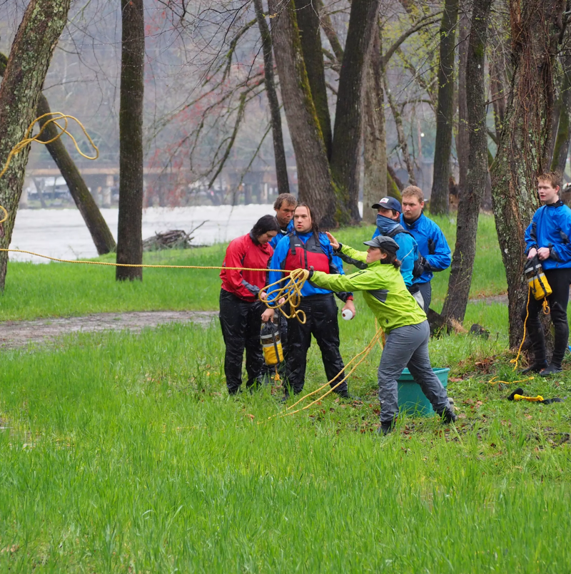 A group of people are standing in a grassy area near trees, possibly a park or outdoor training ground. They appear to be participating in some kind of rescue or team-building exercise, as one person is throwing a rope towards a body of water in the background. Most of them are wearing outdoor clothing, including jackets and hats, suggesting it might be a cool day.